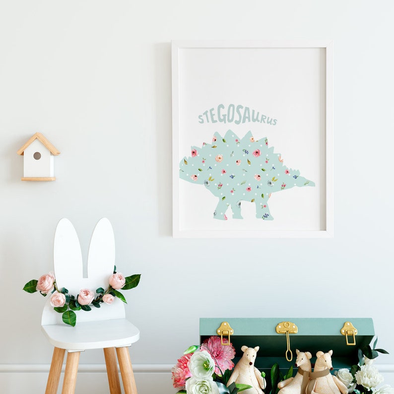 May include: A framed print featuring a light blue Stegosaurus dinosaur silhouette filled with colorful flowers and white polka dots. A white bunny-eared chair with pink flowers and a small wooden birdhouse are also visible.