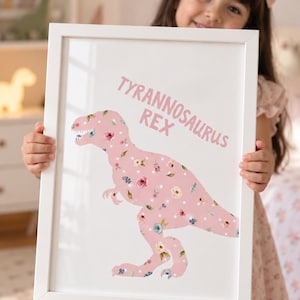 May include: A white rectangular frame containing a print of a Tyrannosaurus Rex silhouette. The dinosaur is light pink with a pattern of white polka dots and various small floral illustrations in shades of pink, blue, and green. The text TYRANNOSAURUS REX is printed in pink above the dinosaur.