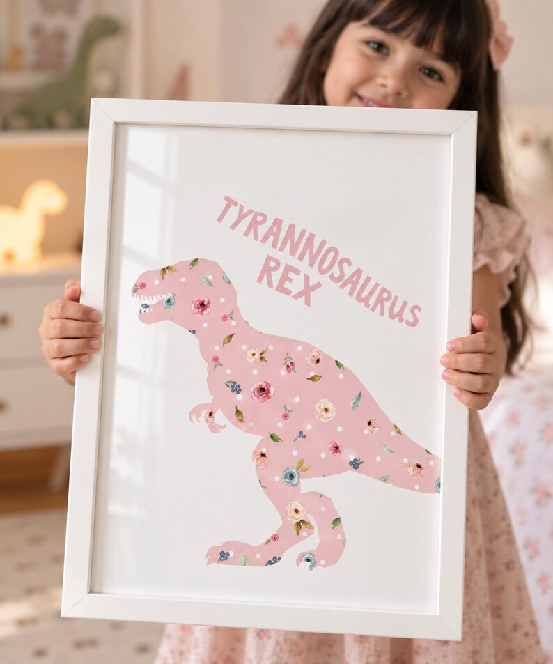 May include: A white rectangular frame containing a print of a Tyrannosaurus Rex silhouette. The dinosaur is light pink with a pattern of white polka dots and various small floral illustrations in shades of pink, blue, and green. The text TYRANNOSAURUS REX is printed in pink above the dinosaur.