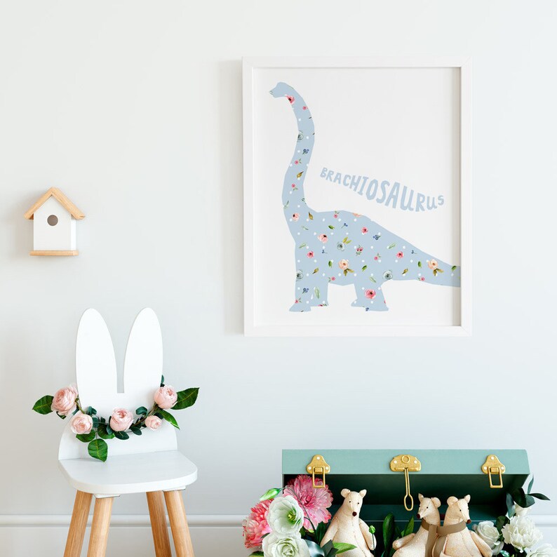 May include: A framed print featuring a light blue Brachiosaurus dinosaur silhouette filled with a floral pattern. The word "BRACHIOSAURUS" is printed in light blue. A small white birdhouse and a white chair with bunny ears and flowers are also visible.