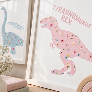 May include: A framed wall art print featuring a pink Tyrannosaurus Rex silhouette filled with a floral and white polka dot pattern. The text TYRANNOSAURUS REX is printed in pink above the dinosaur. A second framed print shows a blue Brachiosaurus.