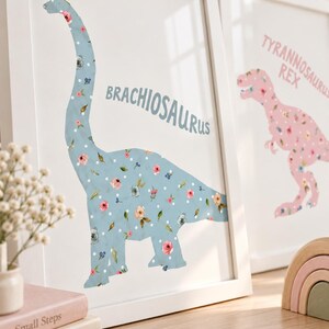 May include: A framed wall art print featuring a light blue Brachiosaurus silhouette with a floral and white polka dot pattern. The text BRACHIOSAURUS is printed in blue. A second framed print shows a pink Tyrannosaurus Rex with similar floral details.