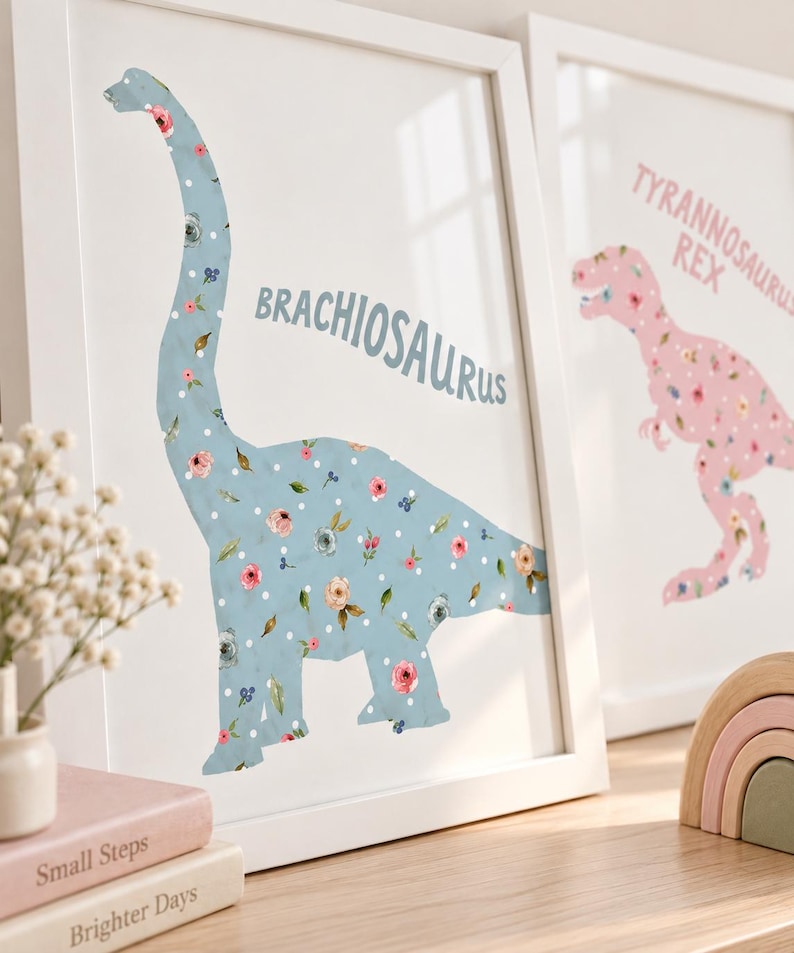 May include: A framed wall art print featuring a light blue Brachiosaurus silhouette with a floral and white polka dot pattern. The text BRACHIOSAURUS is printed in blue. A second framed print shows a pink Tyrannosaurus Rex with similar floral details.