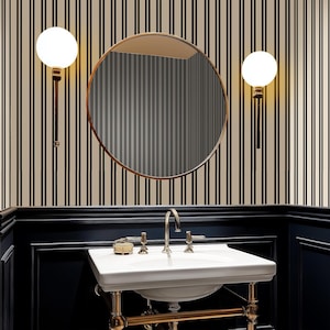 Puede incluir: Interior de baño con espejo redondo con marco de bronce, lavabo blanco con grifería dorada y dos apliques de pared con forma de globo. La pared tiene un patrón de rayas negras y beige, y la pared inferior es azul marino.
