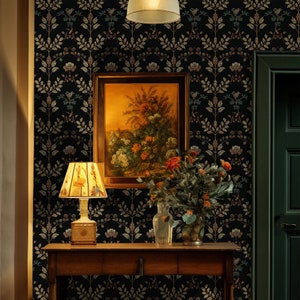 May include: A dark floral wallpaper with a gold and brown pattern. A framed painting of a floral still life hangs on the wall above a wooden table with a lamp and a vase of flowers. A dark green door is to the right of the table.
