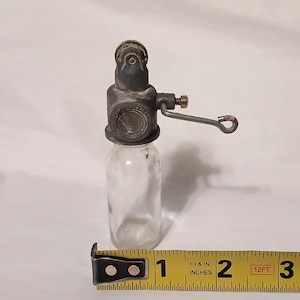 May include: A vintage glass bottle with a grey metal mechanical top featuring a small crank handle and a circular dial. A yellow measuring tape is positioned at the base, showing the object stands approximately 3.5 inches in total height.