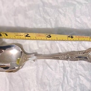 May include: A silver spoon with an ornate handle, approximately 4 inches long, lies next to a yellow measuring tape. The spoon has a rounded bowl and decorative detailing on the handle.