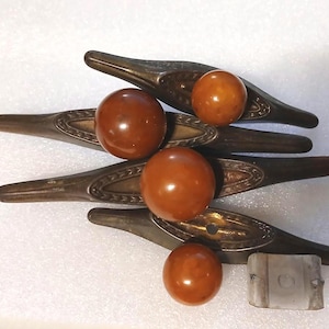 May include: A collection of vintage cabinet hardware, including four dark bronze-toned pulls with decorative detailing and four amber-colored spherical knobs. A small, rectangular, silver-toned piece is also present. The hardware is likely for furniture restoration.