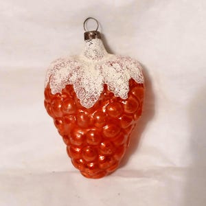 May include: A glass ornament shaped like a strawberry with a shiny orange, textured surface and a white, glittery top. The ornament features a small metal cap at the top with a circular wire loop for hanging, designed with a realistic fruit texture.