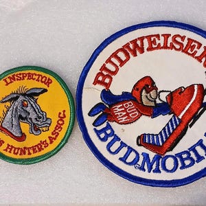 May include: Two embroidered patches. One is round with a yellow background, a gray donkey head, and the text "Wild Ass Hunter's Assoc." The other is a larger, circular patch with "Budweiser Budmobilers" and a cartoon figure.