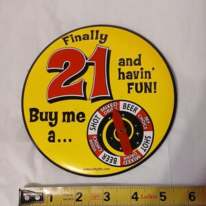 May include: A round yellow game board with the text "Finally 21 and havin' FUN! Buy me a..." and a spinning red arrow over a wheel with drink options. A Lufkin measuring tape shows the item is approximately 6 inches wide at its widest point.