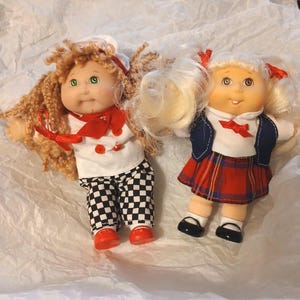 May include: Two vintage Cabbage Patch Kids dolls. One doll is dressed as a chef in a white uniform with a red neckerchief and checkered pants. The other doll wears a plaid skirt, a white shirt, and a blue jacket. Both dolls have blonde hair.