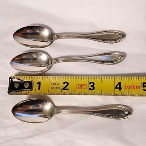 May include: Three silver spoons with ornate handle detailing are displayed next to a yellow measuring tape. The spoons are approximately 4 inches long. The spoons are arranged on a white surface.