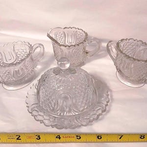 May include: A clear glass serving set with a butter dish, two small handled cups, and a creamer. The set has a textured pattern and is likely vintage. The butter dish is approximately 4 inches in diameter.