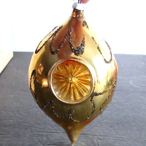 May include: A gold glass Christmas ornament with a black glitter design. The ornament has a round, clear glass center with a starburst pattern.