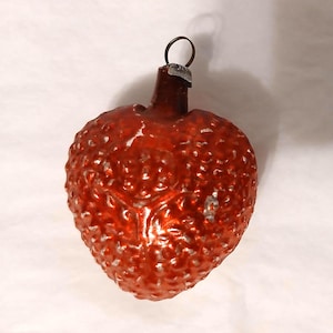 May include: A vintage-style glass Christmas ornament shaped like a strawberry with a textured, bumpy surface in a deep, glossy red finish. The ornament features a small silver-colored metal cap at the top with a thin wire loop for hanging.