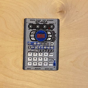 May include: Embroidered patch featuring a gray and black SP-404 sampler. The patch has a rectangular shape with a detailed design of buttons, knobs, and a central circular element with the text "BRKS". Numbers 1-12 are visible.