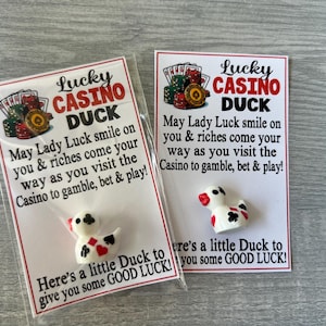 May include: Two "Lucky Casino Duck" novelty gifts. Each features a white duck figurine with black and red accents, packaged with a card that includes casino-themed text and graphics. The cards are in clear plastic.