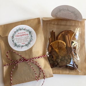 May include: Two brown paper pouches containing dried potpourri ingredients. One pouch is tied with red and white twine and features a circular label reading The Scent of Christmas. The other pouch has a clear window showing dried orange slices, cinnamon sticks, and spices.