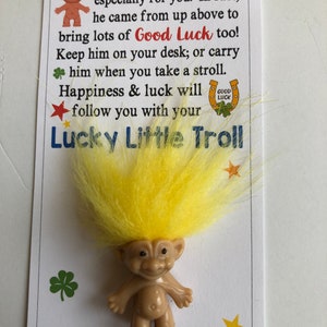 May include: A yellow-haired troll figurine with a friendly expression. The troll is holding a horseshoe with the words "Good Luck" on it. The text on the card reads: "This friendly little Troll is especially for you. In fact, he came from up above to bring lots of Good Luck too! Keep him on your desk; or carry him when you take a stroll. Happiness & luck will follow you with your Lucky Little Troll."