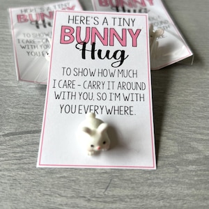 May include: A Tiny Bunny Hug gift set. The set includes a small white bunny figurine and a card with pink and black text that reads "Here's a Tiny Bunny Hug". The card's message is "To show how much I care - carry it around with you, so I'm with you everywhere."