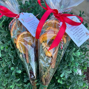May include: Two clear cone-shaped plastic bags filled with dried orange slices, cinnamon sticks, and evergreen sprigs. Each bag is tied with a red ribbon and includes a white tag printed with the text: The scent of Christmas. Add contents to 2 c. water to saucepan & bring to boil, Simmer. Add water as needed & Enjoy!
