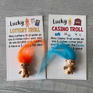 May include: Two novelty troll dolls with colorful hair, one orange and one blue, each on a card. The cards read "Lucky Lottery Troll" and "Lucky Casino Troll" with related graphics and text. The dolls are tan-colored.