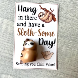 May include: A white card with the text "Hang in there and have a Sloth-Some Day! Sending you Chill Vibes!" features a cartoon sloth and a small sloth figurine. The card has a brown border and is on a gray surface.