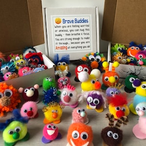 May include: A collection of colorful, fuzzy, handmade toys with googly eyes. The toys are in two open boxes and scattered on a wooden surface. The boxes have a sign that reads "Brave Buddies" and says "When you are feeling worried or anxious, you can hug this buddy - then breathe & focus. You are strong enough to make it through... because you are Amazing at everything you do!"