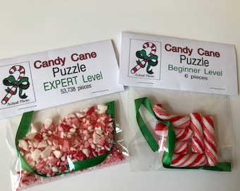 May include: Two candy cane puzzles are displayed. One is labeled "EXPERT Level" with 53,738 pieces, and the other is "Beginner Level" with 6 pieces. Both puzzles feature red and white candy cane pieces in clear plastic bags, tied with green ribbon.