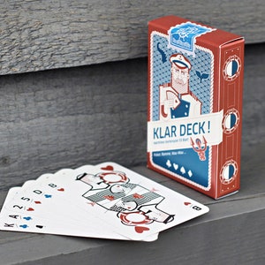 Puede incluir: Una baraja de cartas con tem&aacute;tica n&aacute;utica. La caja presenta una ilustraci&oacute;n de dibujos animados de un marinero con un esquema de color rojo y azul. El texto en la caja dice "Klar Deck!" y "Havet Karmmer til Alt".