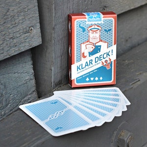 Puede incluir: Una baraja de cartas roja y azul con tem&aacute;tica n&aacute;utica. La caja presenta una ilustraci&oacute;n de dibujos animados de un marinero con barba y un gorro de capit&aacute;n. El texto de la caja dice "Klar Deck!" e incluye los palos de las cartas: "Poker, Romm&eacute;, Mau Mau".