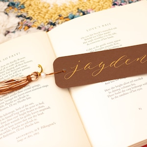 May include: A brown bookmark with a gold tassel and the name "jayden" written in gold calligraphy. The bookmark is lying on an open book with a poem about love.