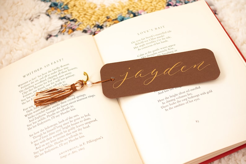 May include: A brown bookmark with a gold tassel and the name "jayden" written in gold calligraphy. The bookmark is lying on an open book with a poem about love.