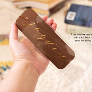 May include: A brown bookmark with a gold inscription that reads "Jayden". The bookmark is in a clear plastic sleeve.