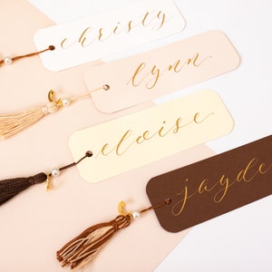 May include: Four personalised bookmarks with gold calligraphy names: "christy", "lynn", "eloise", and "jayden". Each bookmark has a tassel attached with a pearl and gold charm.