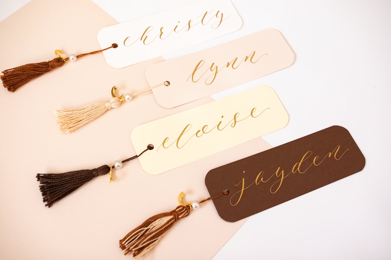 May include: Four personalised bookmarks with gold calligraphy names: "christy", "lynn", "eloise", and "jayden". Each bookmark has a tassel attached with a pearl and gold charm.