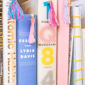 May include: A bookshelf with several books, including "Atomic Habits" by James Clear, "Essays Two" by Lydia Davis, and "1Q84" by Haruki Murakami. Each book has a colourful tassel bookmark.