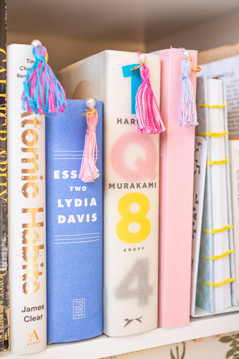 May include: A bookshelf with several books, including "Atomic Habits" by James Clear, "Essays Two" by Lydia Davis, and "1Q84" by Haruki Murakami. Each book has a colourful tassel bookmark.