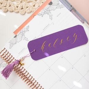 May include: A purple bookmark with the name "Kelsey" written in gold script. The bookmark is placed in a planner with a rose gold spiral binding. The planner has a floral design and a tab labelled "September".