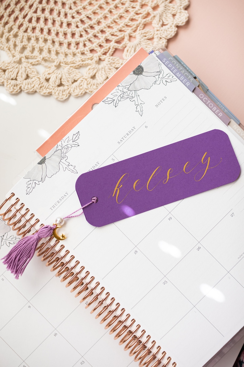 May include: A purple bookmark with the name "Kelsey" written in gold script. The bookmark is placed in a planner with a rose gold spiral binding. The planner has a floral design and a tab labelled "September".