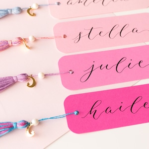 May include: Four pink and white bookmarks with gold crescent moon charms and tassels. The bookmarks are labeled with the names "hailey", "julie", "stella", and an unreadable name.