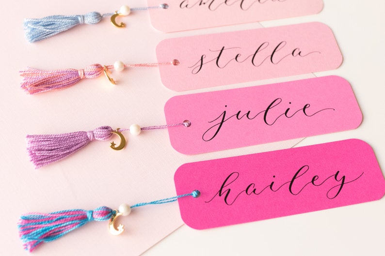 May include: Four pink and white bookmarks with gold crescent moon charms and tassels. The bookmarks are labeled with the names "hailey", "julie", "stella", and an unreadable name.