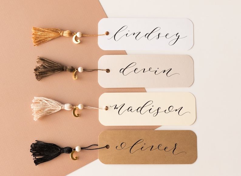May include: Four personalized bookmarks with different colored backgrounds and names written in calligraphy. Each bookmark has a gold crescent moon charm and a tassel.