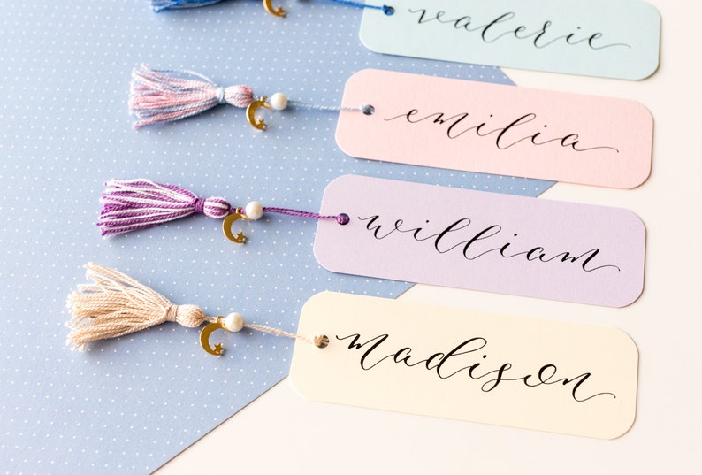 May include: Four colorful bookmarks with gold crescent moon charms and tassels. The bookmarks are labeled "Valerie", "Emilia", "William", and "Madison".