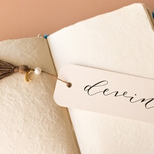 May include: A white bookmark with the word "devin" written in black calligraphy. The bookmark is attached to a brown tassel with a gold charm and a white pearl.