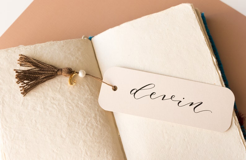 May include: A white bookmark with the word "devin" written in black calligraphy. The bookmark is attached to a brown tassel with a gold charm and a white pearl.