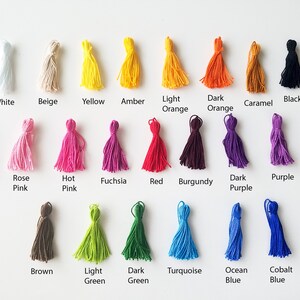 Peut inclure: Une collection de 20 pompons color&eacute;s dans diff&eacute;rentes nuances de blanc, beige, jaune, orange, rouge, rose, violet, marron, vert, bleu et noir. Les pompons sont dispos&eacute;s en rang&eacute;es sur une surface blanche.
