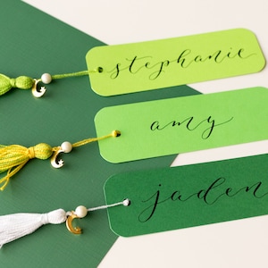 May include: Three green bookmarks with gold crescent moon charms and tassels. The bookmarks are labeled "Stephanie", "Amy", and "Jaden" in black calligraphy.