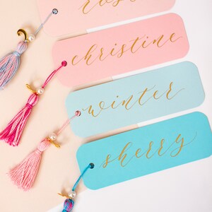 May include: Four colourful bookmarks with gold lettering and tassels. The bookmarks are pink, light blue, and blue. The names "Reese", "Christine", "Winter", and "Sherry" are written in gold calligraphy on the bookmarks.